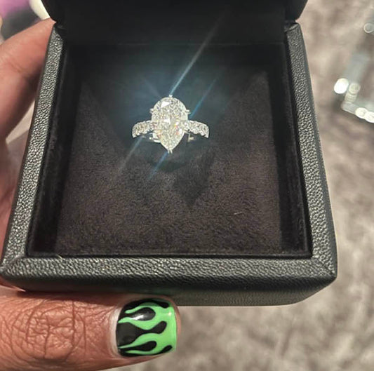 Close-up of a diamond engagement ring in a black velvet box, with a sparkling oval-shaped center stone and diamond accents, held by a hand with green flame nail art.