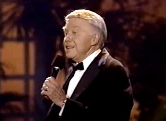 Legendary entertainer performing on stage, dressed in a tuxedo, holding a microphone and smiling.