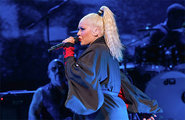 Gwen Stefani performing live on stage, wearing a black outfit with a high ponytail, holding a microphone with a dramatic backdrop of the band playing.