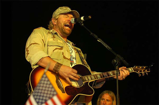 Toby Keith performing live on stage, singing passionately into the microphone while playing guitar, with an American flag visible in the foreground.