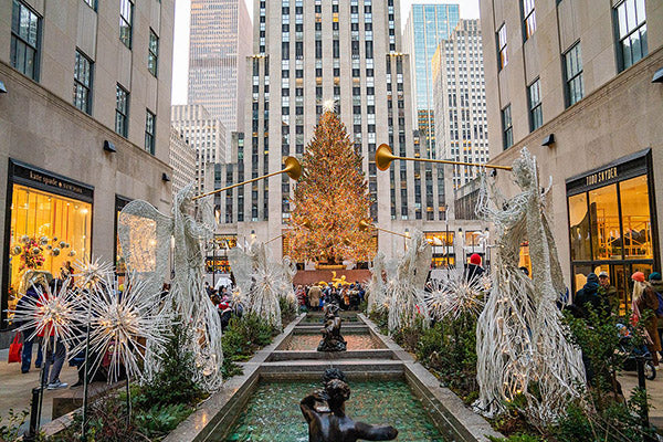 Rockefeller Center Christmas tree surrounded by festive lights and angel sculptures in New York City, with people admiring the holiday decorations.