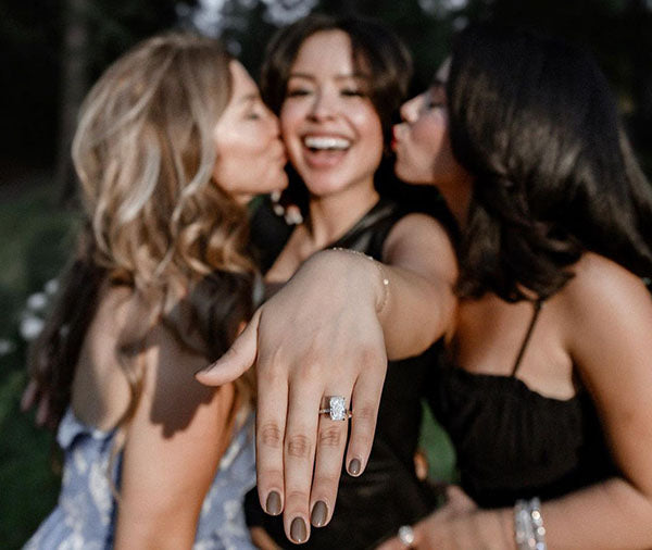 Three women celebrating engagement, with one showing off her large diamond ring, smiling and posing outdoors.