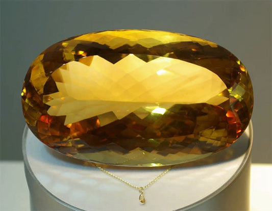 Large, oval-shaped yellow gemstone with a brilliant cut, displayed on a pedestal with a golden necklace in the background.
