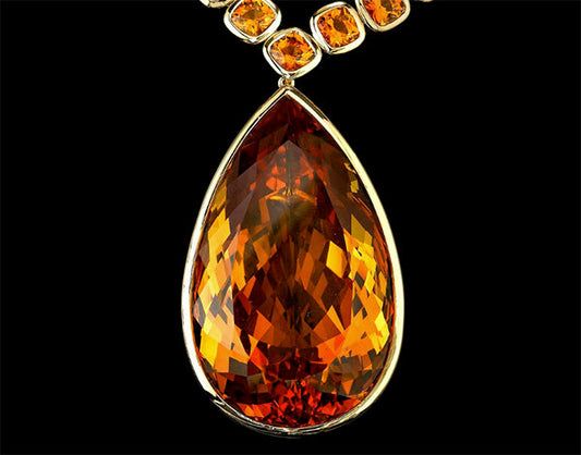 Close-up of a luxurious gold necklace featuring a large pear-shaped orange gemstone at the center, surrounded by smaller round orange gemstones. The pendant shines brightly against a black background.