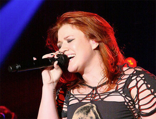 Kelly Clarkson performing live on stage, singing passionately into a microphone with vibrant lighting highlighting her red hair and energetic expression.