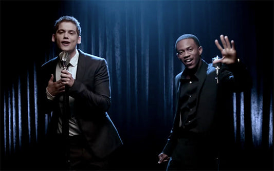 Two male singers performing on stage with microphones, dressed in formal suits, under spotlight with dramatic lighting.