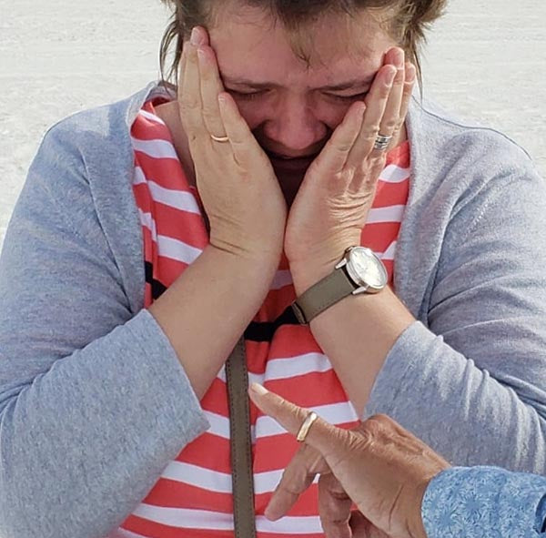 A woman in a striped shirt and gray cardigan, visibly emotional, covers her face with her hands while a hand points at her, possibly marking a moment of surprise or proposal.
