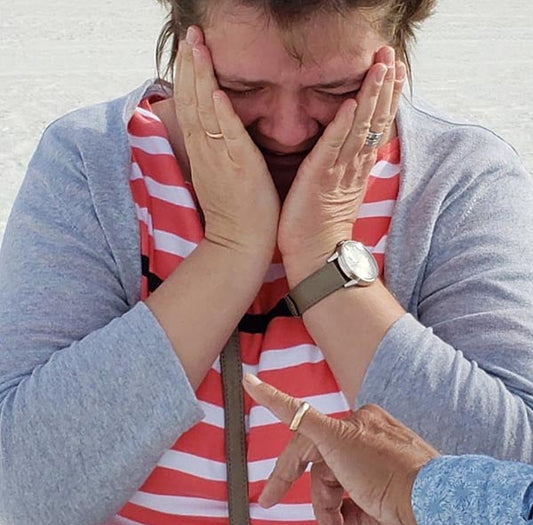 A woman in a striped shirt and gray cardigan, visibly emotional, covers her face with her hands while a hand points at her, possibly marking a moment of surprise or proposal.