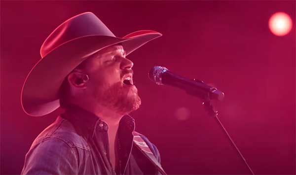 Country music singer wearing a cowboy hat, passionately performing with a guitar and microphone, captured in vibrant stage lighting during a live concert performance.
