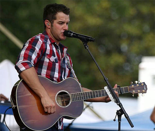 Male country music singer performing live on stage with an acoustic guitar. He is wearing a red and white checkered shirt and a microphone headset, captured in a vibrant outdoor concert setting.