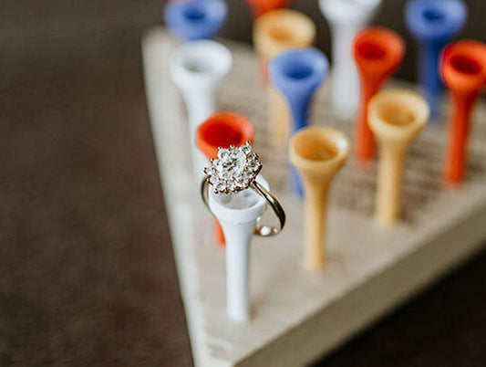A sparkling diamond engagement ring placed on a white golf tee, with colorful golf tees in the background, symbolizing a unique and creative engagement moment.