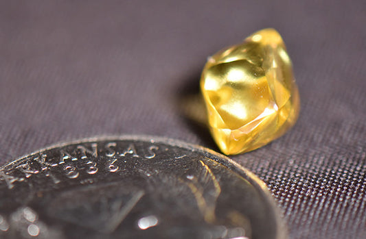 Close-up of a yellow gemstone placed next to a silver coin, with intricate details visible on both the gemstone and the coin. The coin displays the word 'Arkansas' and the year '1936.