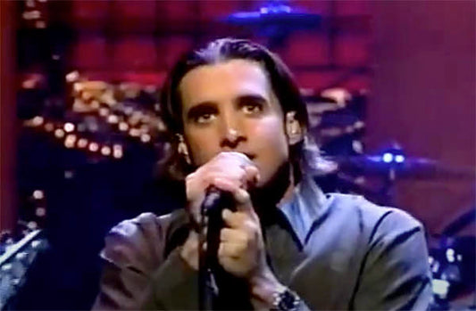 Scott Stapp of Creed performing live on stage, holding a microphone and singing passionately. The image captures his intense expression during the performance, with a blurred band setup in the background.