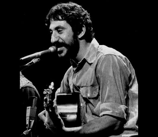 Black and white image of a musician playing guitar and singing into a microphone. The man has a beard and mustache and is wearing a button-up shirt, performing on stage.