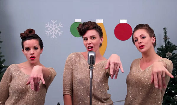 Three women singing into a vintage microphone, wearing sparkling gold dresses with Christmas decorations in the background.