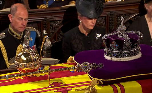Royal funeral scene with the Crown Jewels, including the Imperial State Crown and Sovereign's Orb, placed on a flag-covered coffin, while mourners are seated in the background.