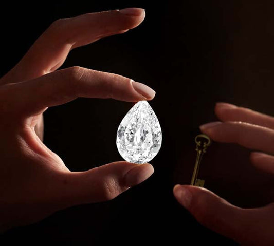 Close-up of a hand holding a large, sparkling pear-shaped diamond with a key in the other hand, set against a dark background.