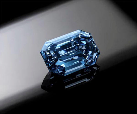 Close-up of a blue sapphire gemstone with an emerald cut, showcasing its vibrant color and sharp facets. The gem is placed on a reflective surface, highlighting its brilliance and clarity.