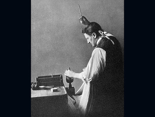 Black and white historical image of a man working with a hammer and chisel, carving or shaping material, with another hand raised above him holding a tool.