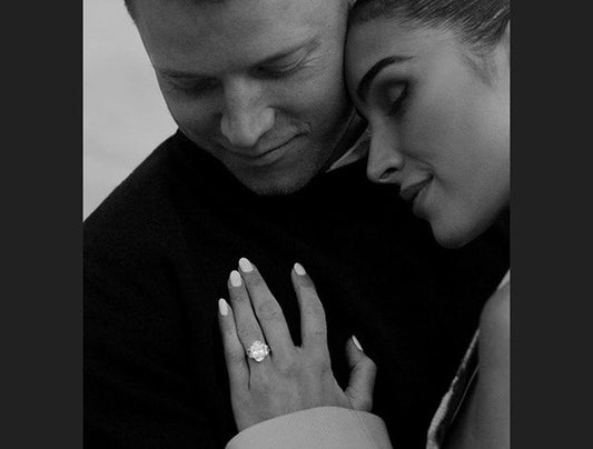Black and white photo of a couple embracing, showcasing an engagement ring on the woman’s hand as she rests her head on her partner’s chest.