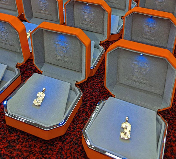 Close-up image of multiple orange jewelry boxes with illuminated interiors, each holding a silver pendant featuring the number 3. The luxurious presentation highlights the exclusive jewelry items.