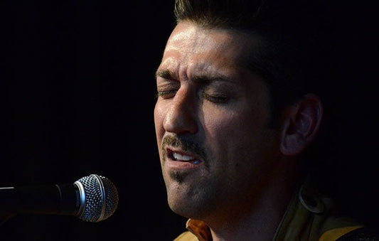 Close-up of a male musician with a mustache singing into a microphone on stage, showing a focused expression while performing live.