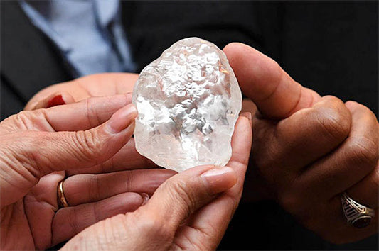 Two hands holding a large, rough diamond with clear, translucent facets, showcasing the raw crystal's natural beauty.