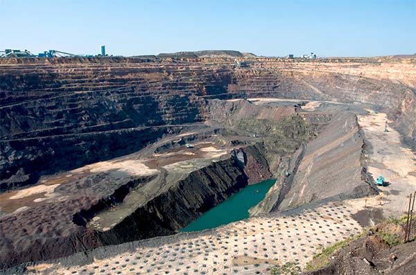 Wide view of a large open-pit mining operation with layers of rock, mining machinery, and a small body of water in the center.