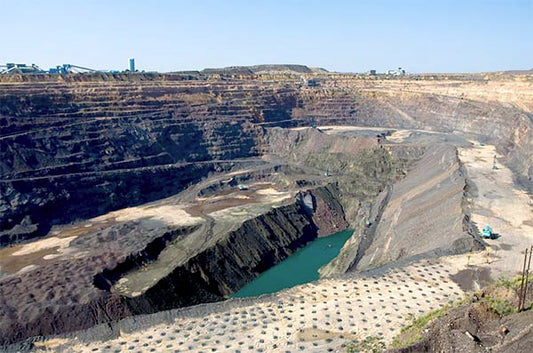 Wide view of a large open-pit mining operation with layers of rock, mining machinery, and a small body of water in the center.