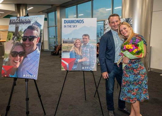 A couple posing for a photo at an airport with large Delta Airlines promotional posters behind them, featuring the words 'Togetherness on Delta Flight' and 'Diamonds in the Sky.' The woman is holding a bouquet of flowers while both smile happily.