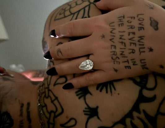 Close-up of a hand with a large pear-shaped diamond engagement ring, tattooed arm with the phrase 'LOVE WILL LIVE FOREVER IN THE INFINITE UNIVERSE' visible.