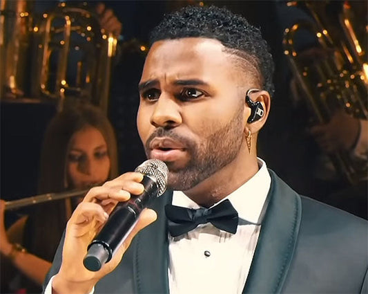 Jason Derulo performing on stage with a microphone, wearing a tuxedo, singing passionately with a live band in the background.