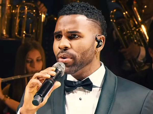 Jason Derulo performing on stage wearing a tuxedo while holding a microphone, with a live orchestra in the background.