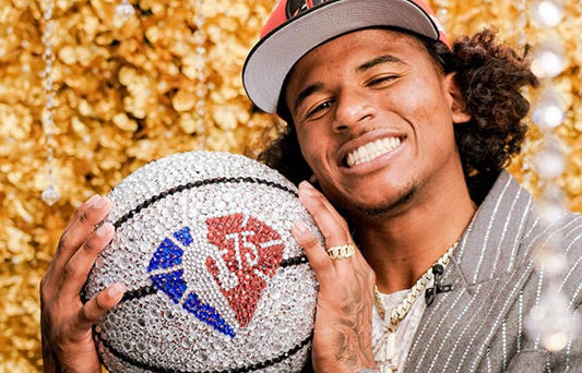 A man smiling while holding a custom diamond-studded basketball featuring a logo, wearing a cap and jewelry, with a gold glittery background.