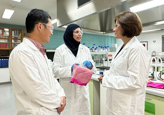Three scientists in lab coats discussing research in a laboratory, one holding a pink cloth, with laboratory equipment in the background.