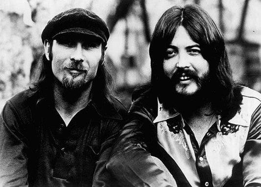 Black and white photo of two musicians sitting together, one wearing a black cap and the other with long hair and a patterned shirt, representing a classic vintage rock band vibe.