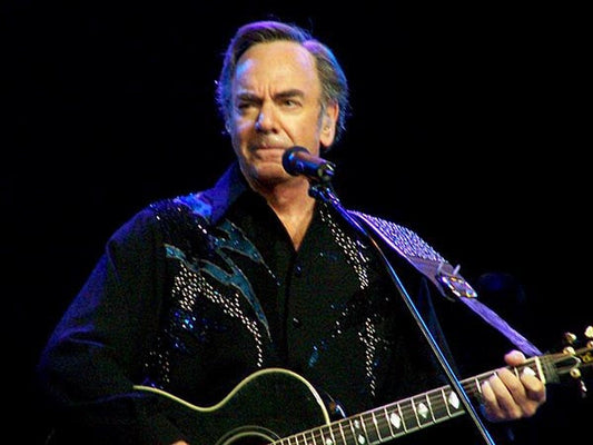 Neil Diamond performing on stage, playing an acoustic guitar and singing, wearing a black shirt with sparkling details.