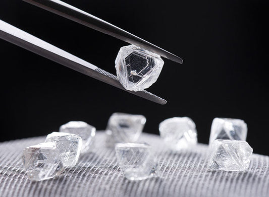 Close-up of a diamond being held with tweezers, surrounded by several rough, uncut diamonds on a dark background, showcasing their raw form and clarity.