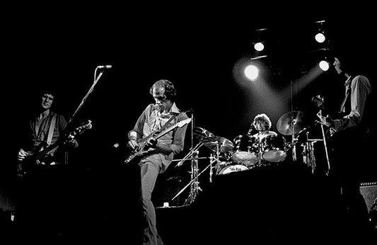 Black and white photo of a rock band performing on stage with a guitarist playing and a drummer in the background under bright stage lights.