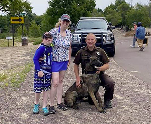 Family posing with a police officer and a K9 dog near a police vehicle at a park.