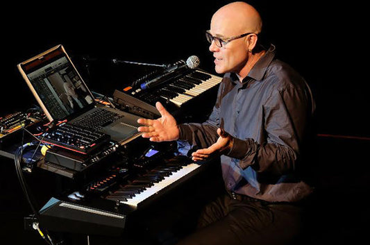 Musician performing live with a keyboard and laptop, using electronic instruments and speaking into a microphone during a stage performance.