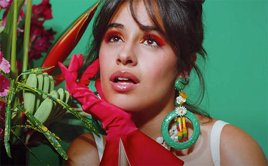 Camila Cabello posing in a colorful fashion shoot, wearing bold red gloves, oversized earrings, and surrounded by vibrant flowers, showcasing her glamorous makeup and striking expression.