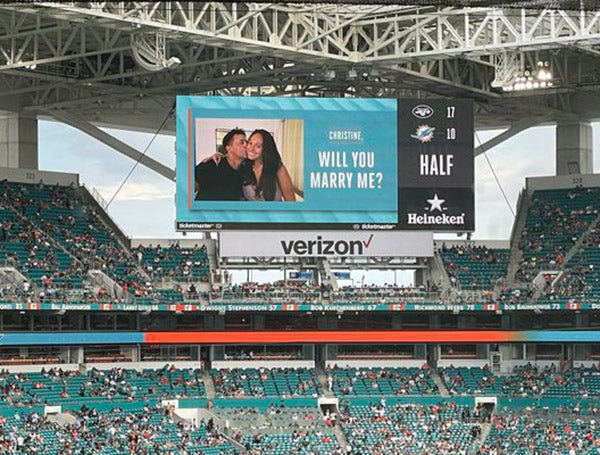 Proposal on stadium Jumbotron screen asking 'Christine, will you marry me?' during a sports game with fans in the background.