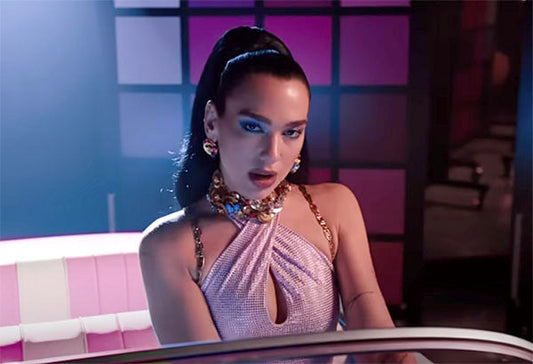 Dua Lipa performing in a glamorous music video, wearing a sparkling purple halter dress, bold jewelry, and sitting in a retro car with pink and purple lighting.