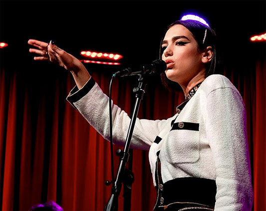 Dua Lipa performing live on stage, holding a microphone with one hand, wearing a chic white and black outfit, under vibrant red stage lights.