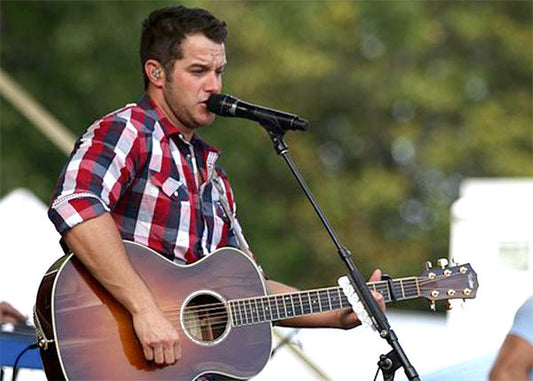 Male country singer performing live on stage with an acoustic guitar, wearing a red plaid shirt and microphone headset.