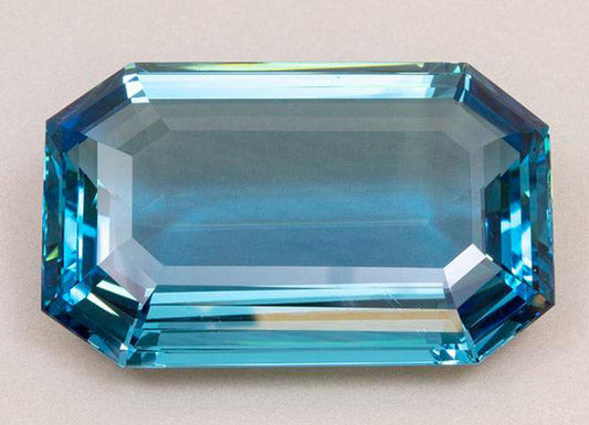 Close-up of a large, rectangular blue topaz gemstone with an octagonal cut. The stone has a brilliant blue color, showcasing its clarity and intricate facets.