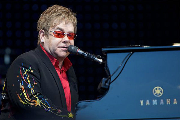 Elton John performing on stage at a Yamaha piano, wearing sunglasses and a red shirt with colorful embroidered jacket.