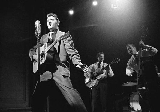 Black and white photo of Elvis Presley performing live on stage with a guitar, accompanied by his band members on bass and drums.