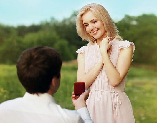 Man proposing with an engagement ring to a smiling woman in a pink dress, in a romantic outdoor setting with a green background.